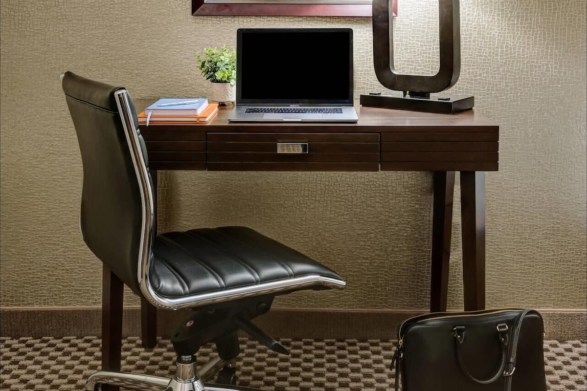 Une chambre d’hôtel équipée d’un bureau, d’une chaise, d’un ordinateur portable, d’un tableau encadré, d’une lampe et d’un sac à main.