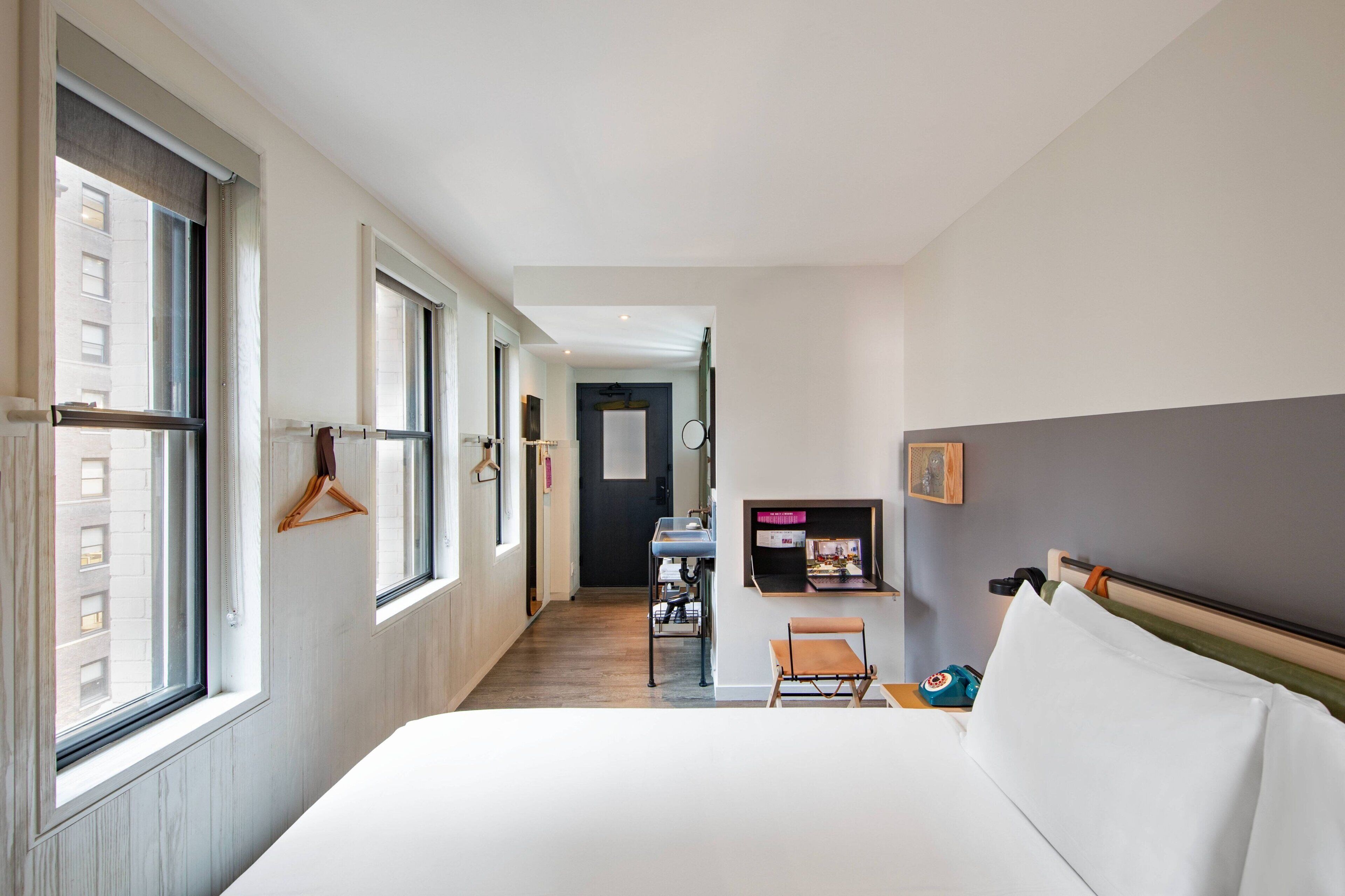 A hotel room with a large bed, a desk, and a chair. There is a window on the left side and a door at the end of the room.