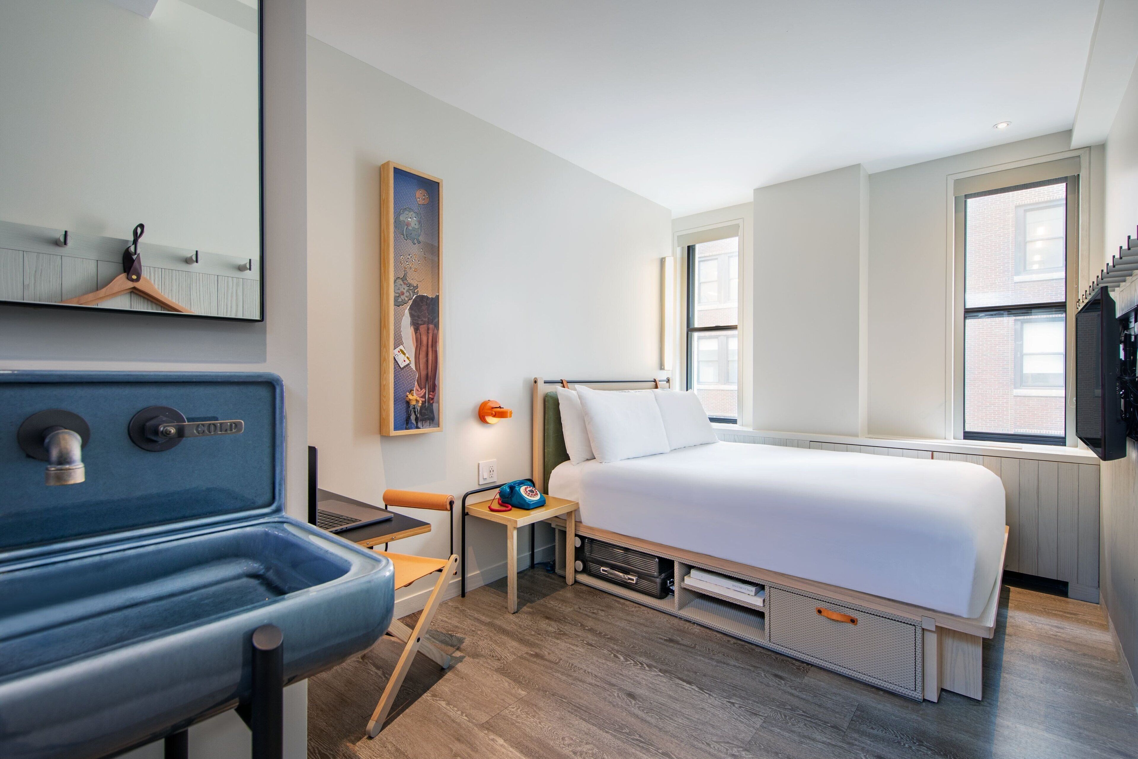 Une chambre d’hôtel moderne, équipée d’un lit, d’un petit bureau avec une chaise, d’un lavabo et d’une grande fenêtre.