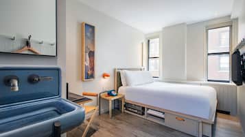 Une chambre d’hôtel moderne, équipée d’un lit, d’un petit bureau avec une chaise, d’un lavabo et d’une grande fenêtre.