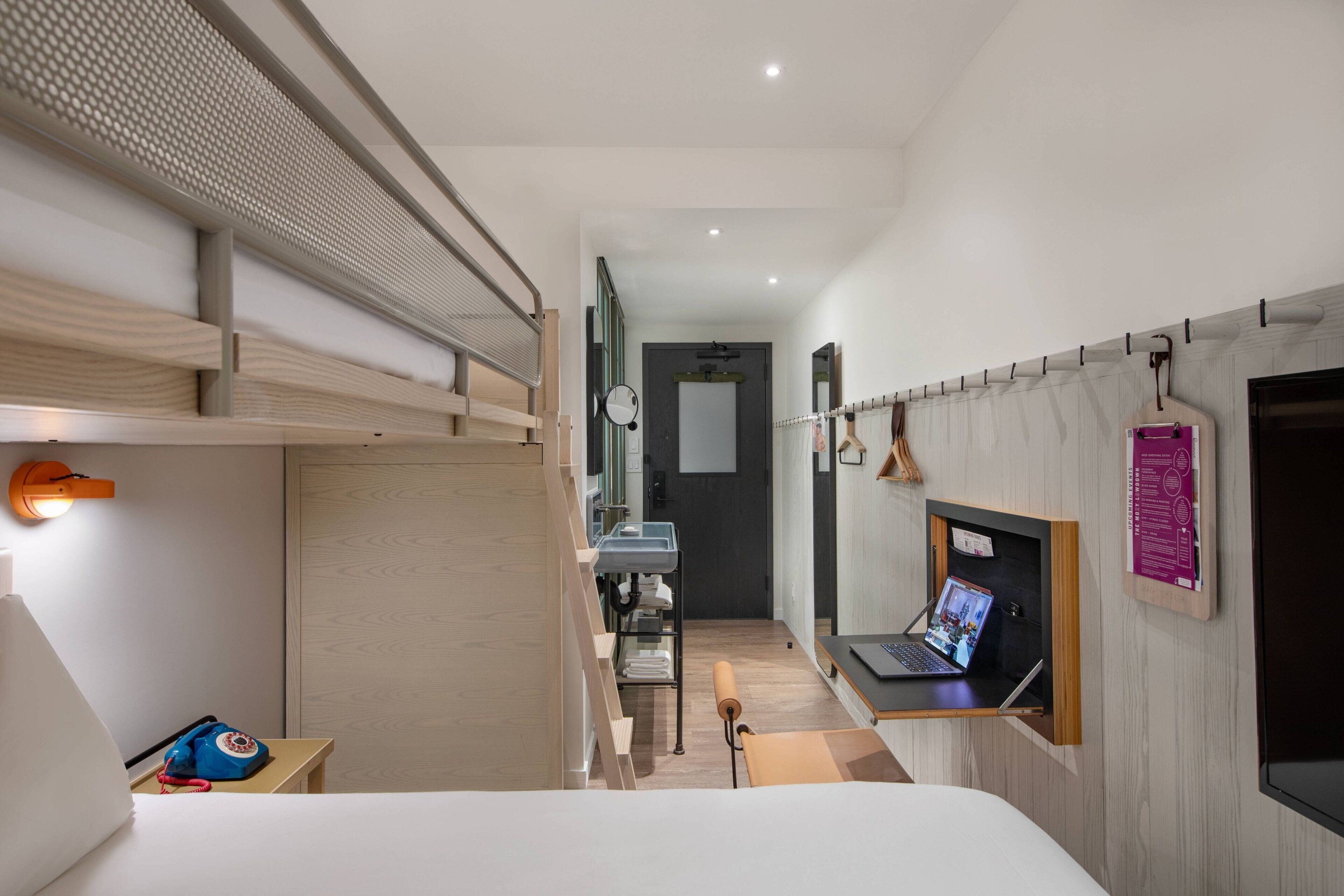 A modern hotel room with a bunk bed, a desk with a laptop, a small table with a telephone, and a wall-mounted TV.