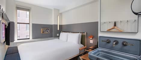 Quarto de hotel com cama, escrivaninha com pia, espelho e janela com vista para edifícios.