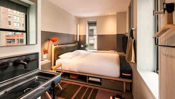A compact hotel room with a bed, a small desk with a lamp, a window with a view of buildings, and a built-in oven.