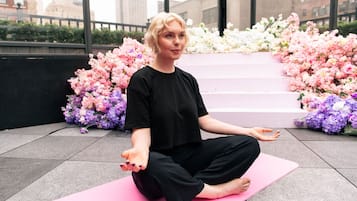 A person sitting on a pink yoga mat outdoors with floral arrangements in the background.