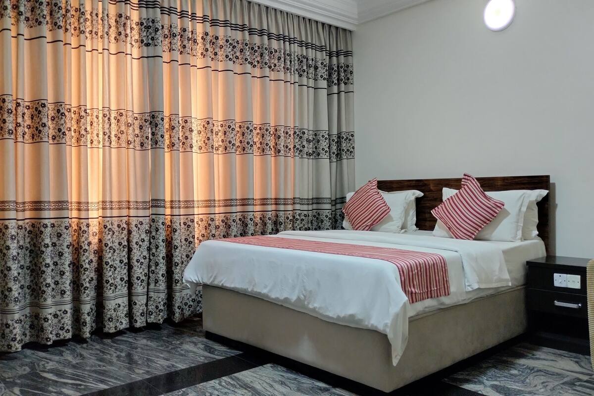 A neatly made bed with white bedding and red striped pillows, a wooden headboard, a nightstand with a lamp, and patterned curtains.