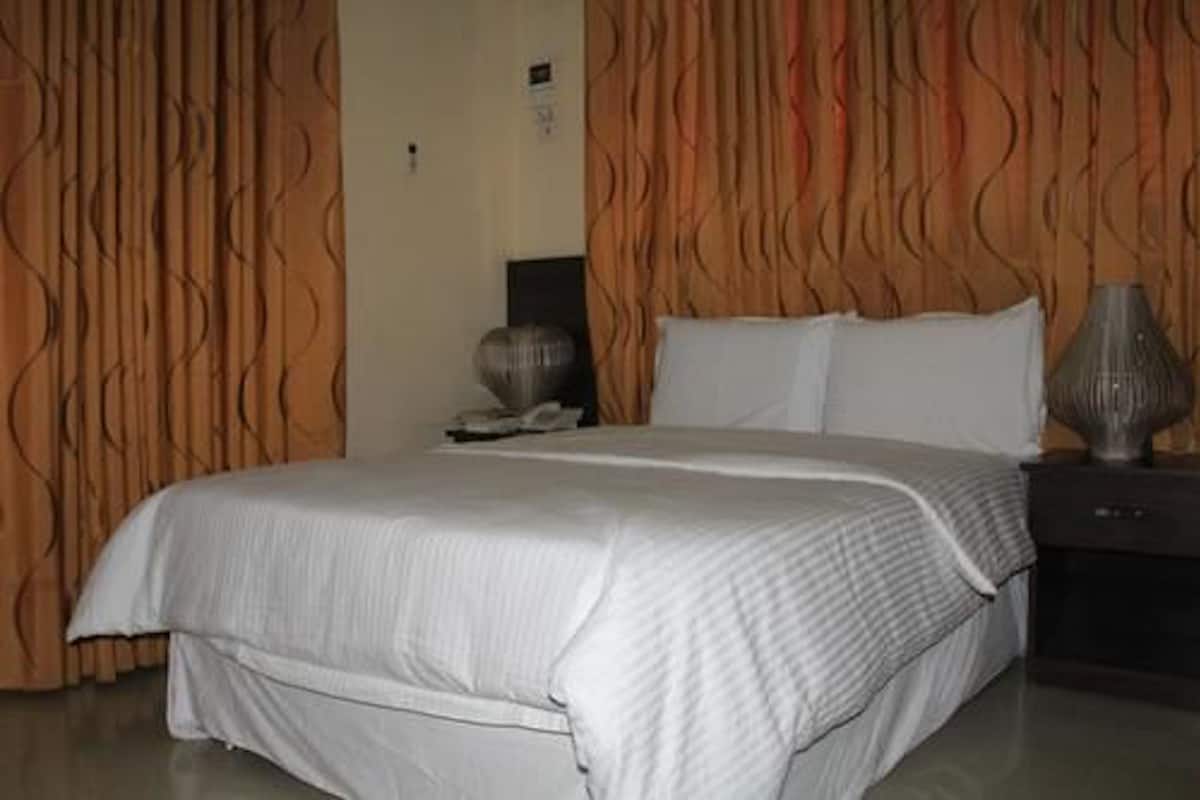 A neatly made bed with white bedding, a dark nightstand with a lamp, and a wall with a wavy pattern.