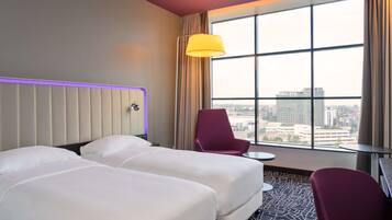 Une chambre d’hôtel avec deux lits, un fauteuil violet, une petite table et une grande fenêtre donnant sur la ville.