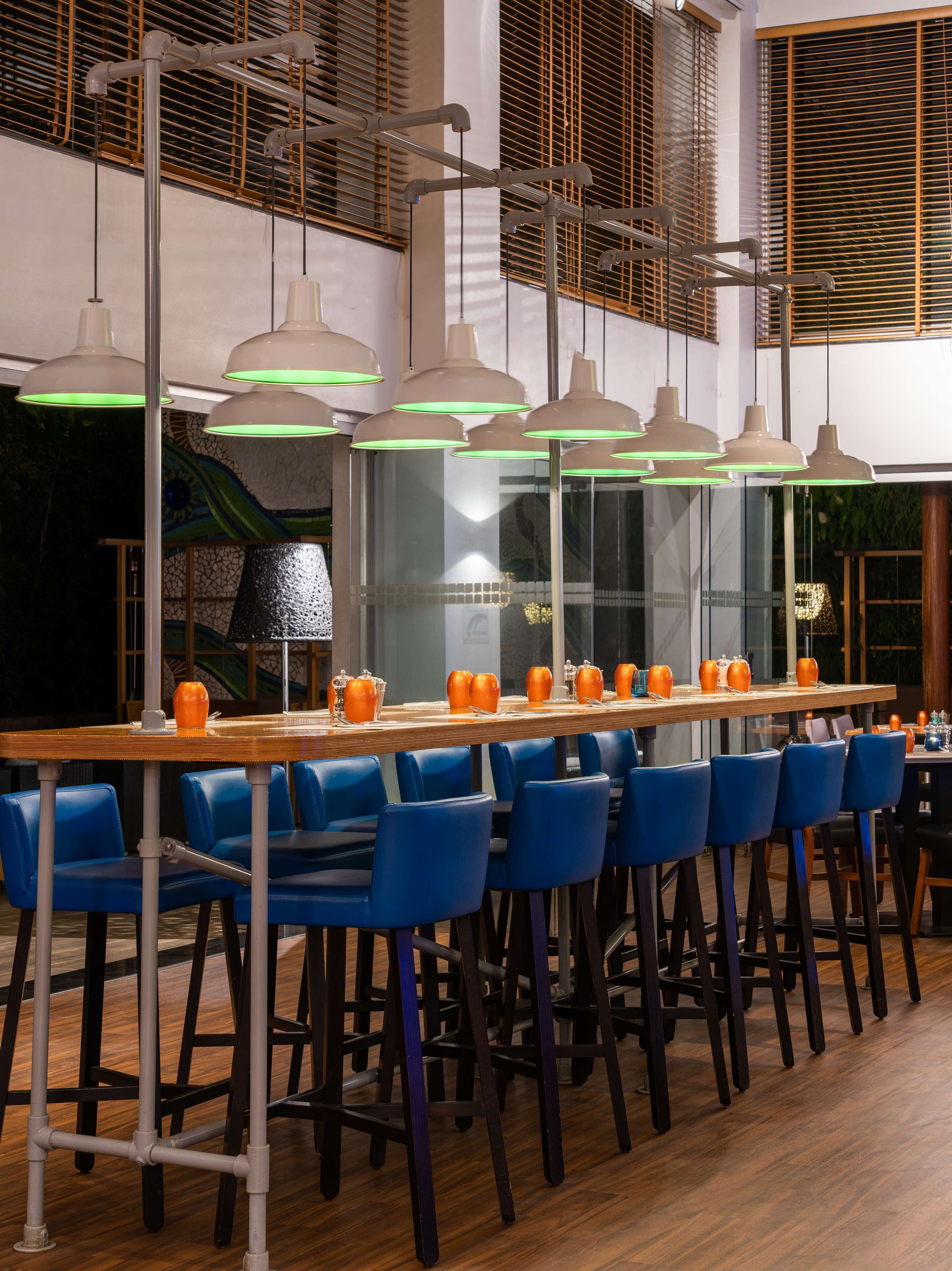 A modern bar area with multiple hanging pendant lights, a long wooden table with orange teapots, and blue chairs.