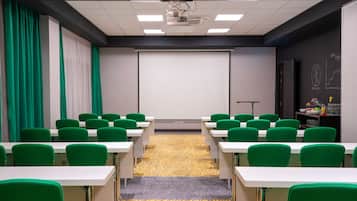 Sala de conferência com cadeiras verdes e mesas brancas, uma tela de projetor e um quadro negro com diagramas.