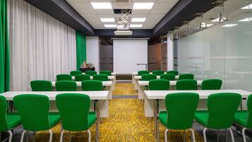 Uma sala de conferências com cadeiras verdes e mesas brancas, uma tela de projetor e um tapete estampado.