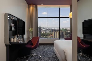 A hotel room with a large window, a bed, two red chairs, a desk with a TV and a coffee maker, and a city view.