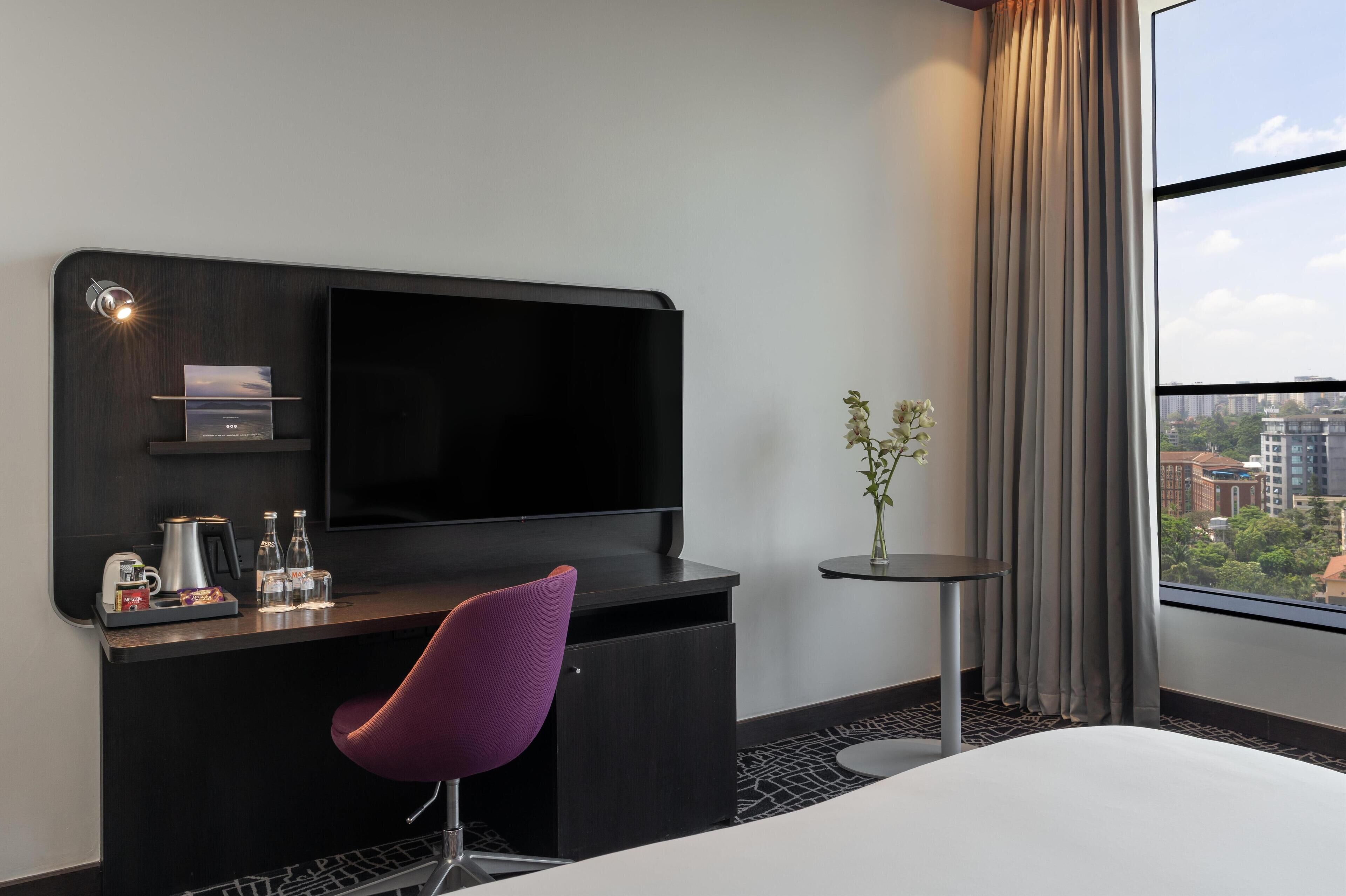 Een hotelkamer met een groot bed, een bureau met een flatscreen televisie, een paarse stoel, een tafeltje met een vaas en uitzicht op de stad door grote ramen.