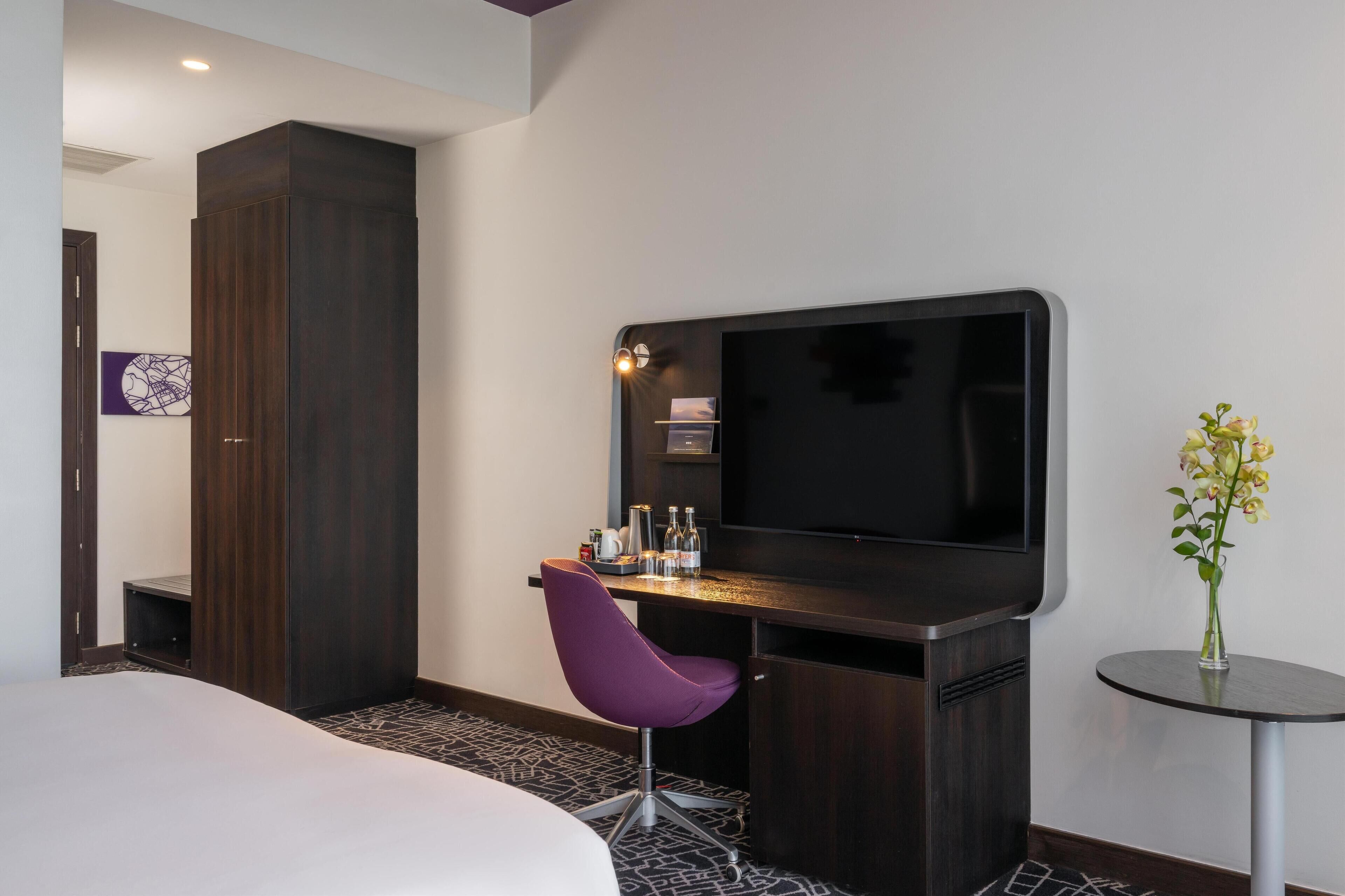 A hotel room with a bed, a desk with a chair, a television, a small table with a vase, and a door leading to another room.