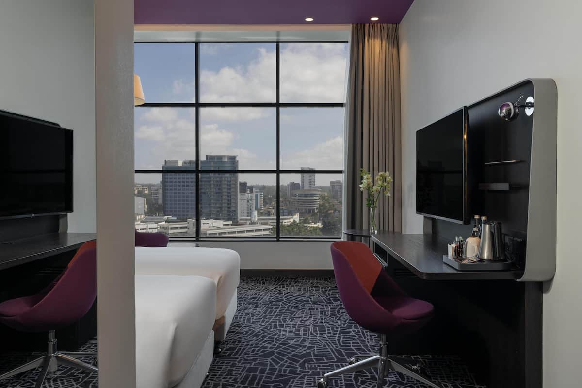 Une chambre d’hôtel comprenant un lit, deux fauteuils, un bureau avec une télévision et une machine à café, ainsi qu’une grande fenêtre donnant sur la ville.