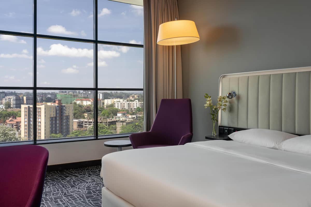 Une chambre d’hôtel avec une grande fenêtre, un lit, un fauteuil violet, une petite table et une table de chevet avec une lampe.