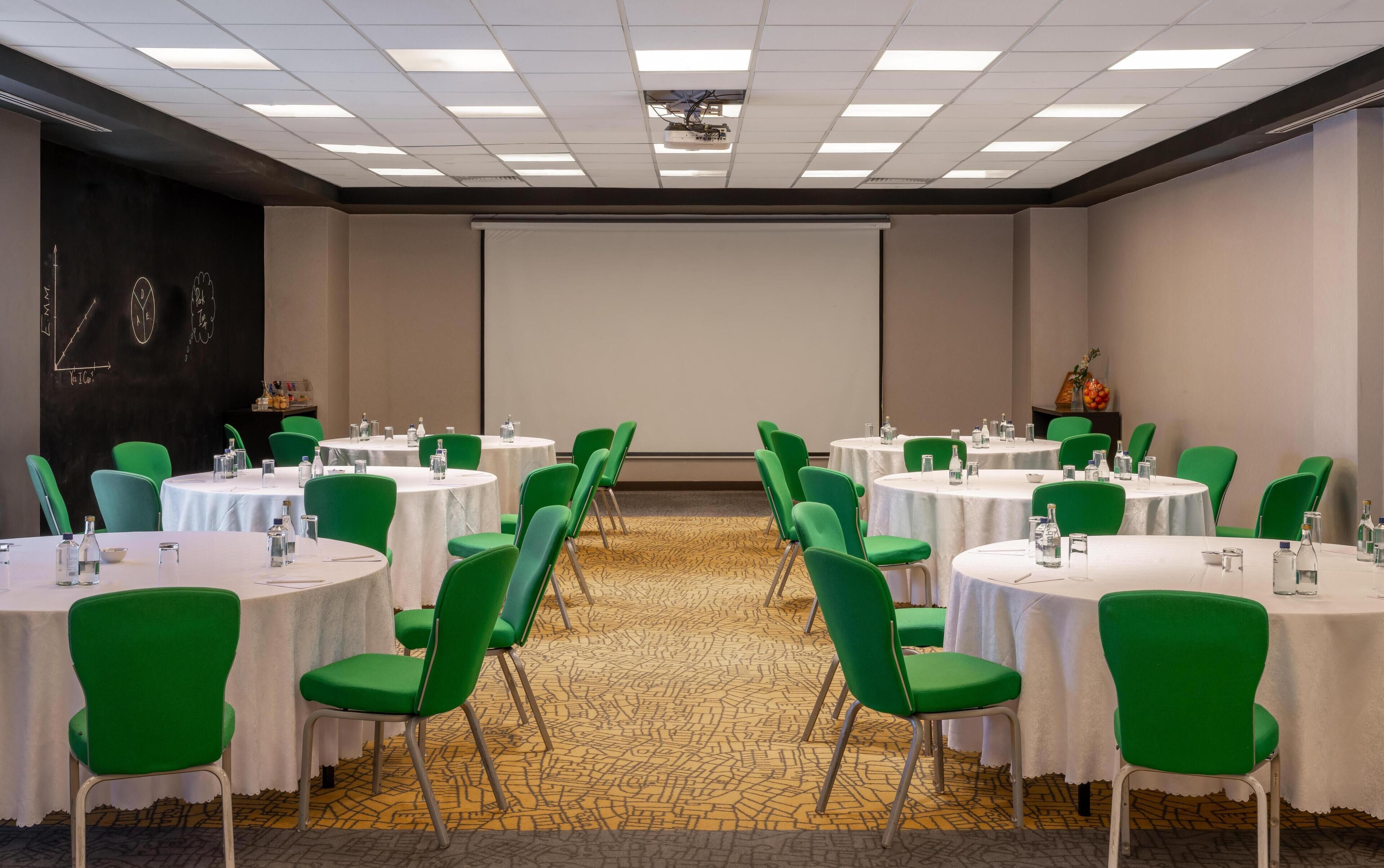 A conference room with round tables, green chairs, and a projector screen.