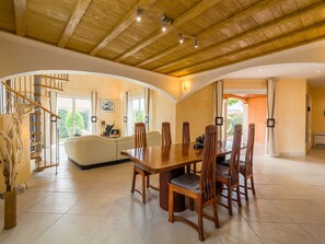 Relax in a spacious room with a high ceiling, dining area, and comfortable seating, plus a staircase to an upper level with views of the surrounding greenery.