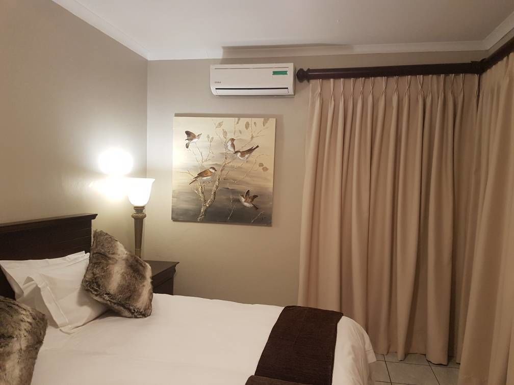 A hotel room with a bed, a nightstand, a lamp, an air conditioning unit, a painting, and a window with curtains.