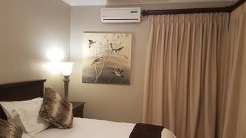 A hotel room with a bed, a nightstand, a lamp, an air conditioning unit, a painting, and a window with curtains.