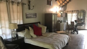 A bedroom with a large bed, a nightstand with a lamp, a wardrobe, a dining table with chairs, and a kitchen area in the background. - Benka Lifestyle (Lobamba)