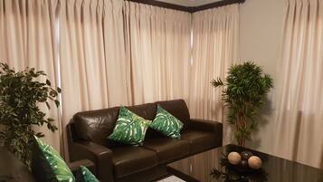 A living room with a brown leather sofa, two green patterned cushions, a glass coffee table, and two potted plants.