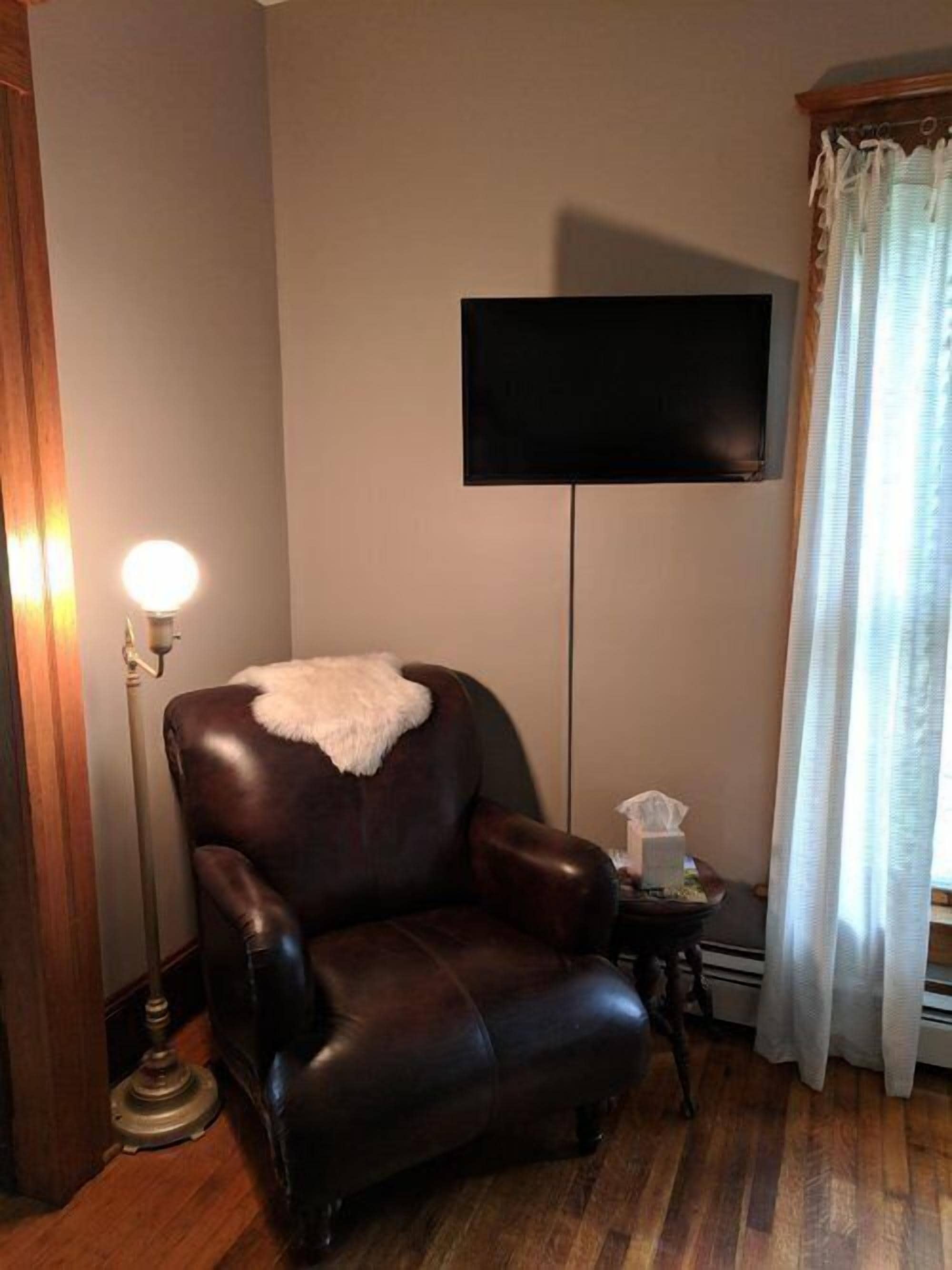A room with a leather armchair, a flat-screen TV mounted on the wall, a floor lamp, and a window with curtains.