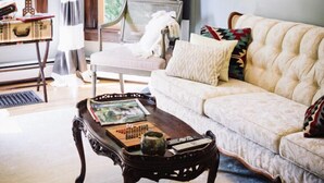 A living room with a tufted sofa, a wooden coffee table with books, a wicker chair, and a view of greenery through large windows. - Burdett House Bed & Breakfast (Burdett)
