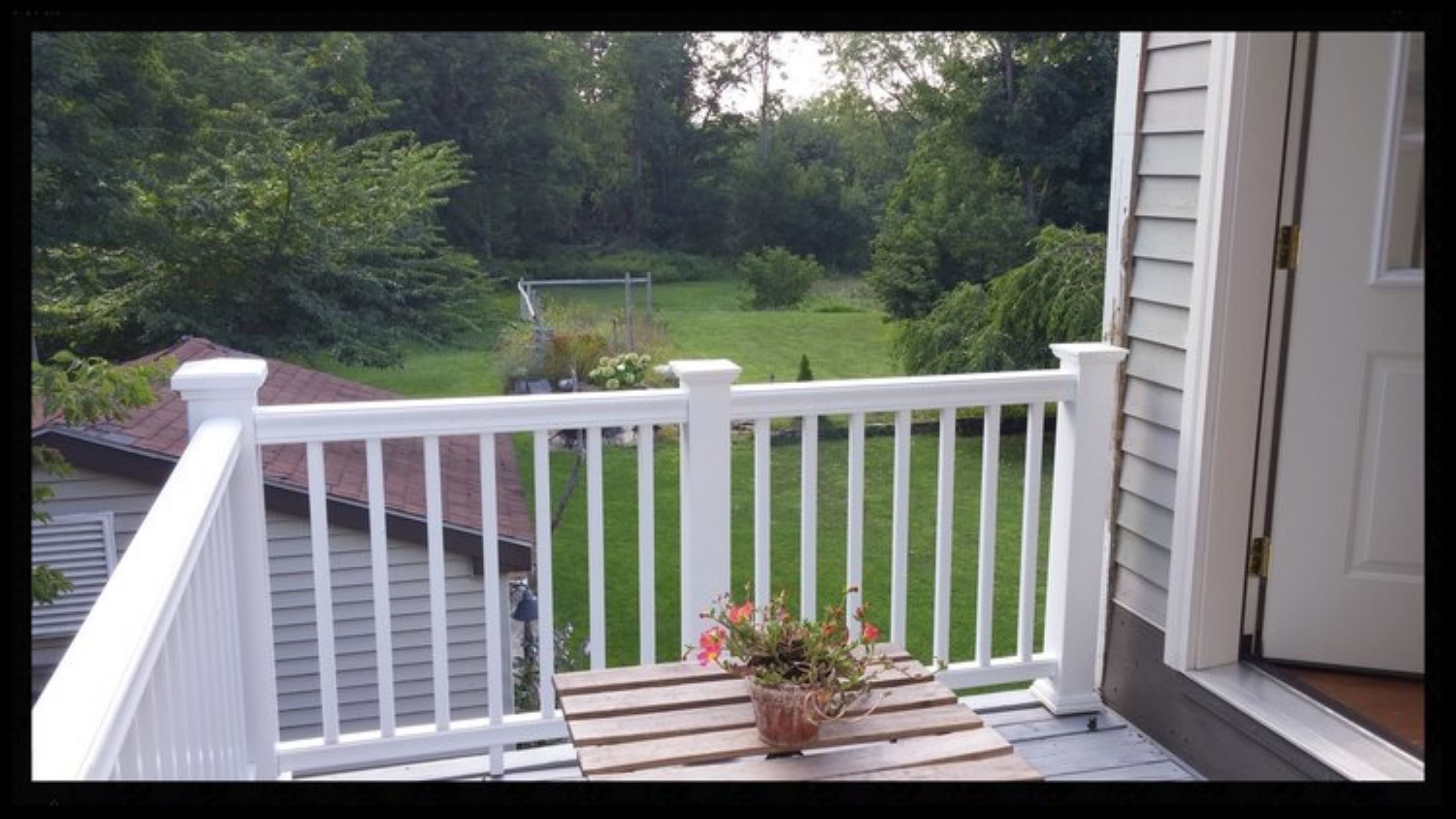 A porch with a white railing, a potted plant, and a view of a green lawn and trees.