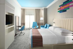 A hotel room with a bed, a desk with a chair, a TV mounted on the wall, and a window with curtains.