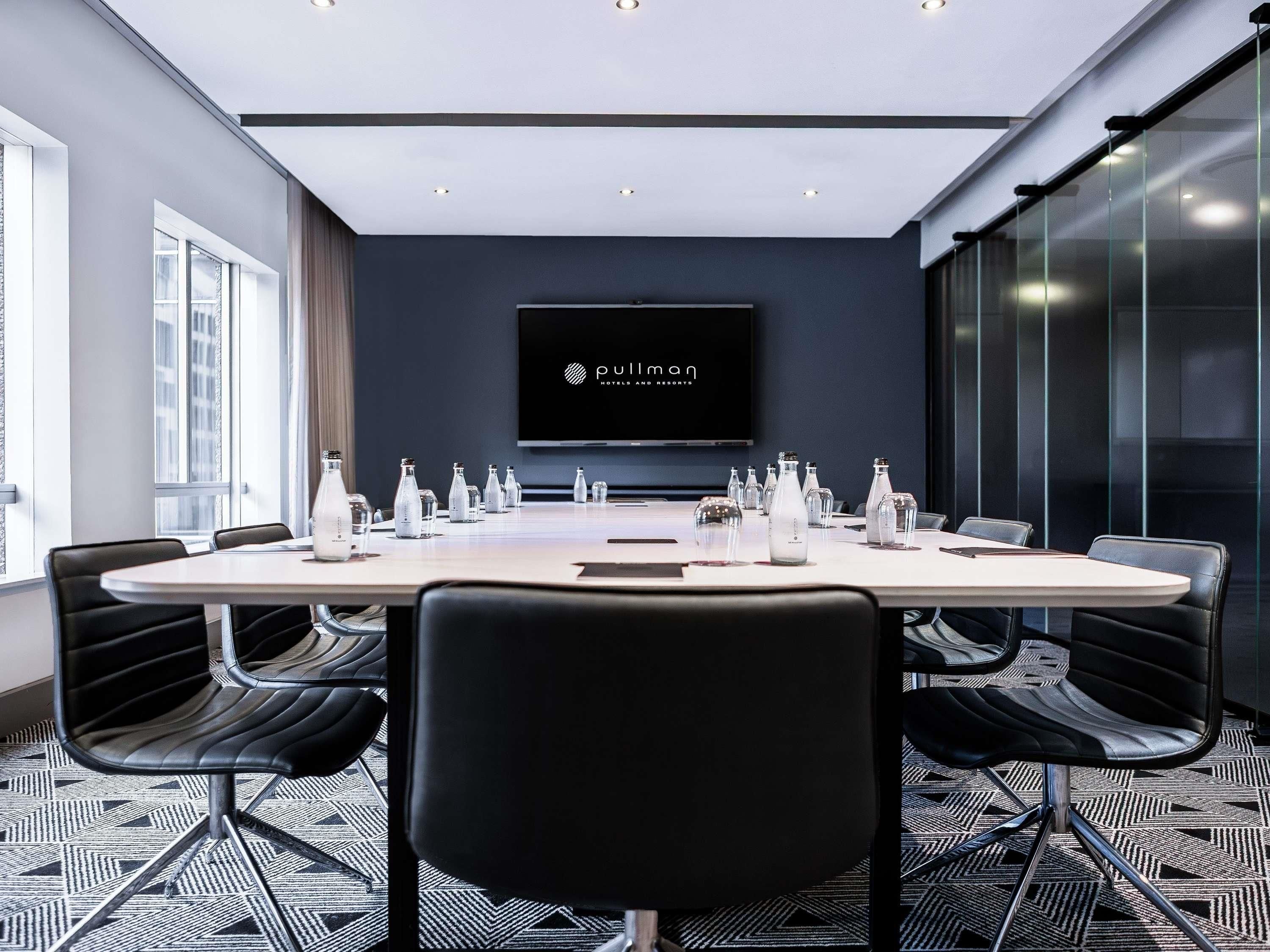 Une salle de conférence avec une longue table, des chaises noires et un écran de télévision affichant le logo « Pullman Hotels & Resorts ».