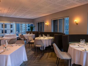 A restaurant with round tables, chairs, and a view of buildings through large windows. - Pullman Cape Town City Centre (Cape Town)