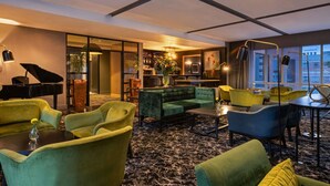 Un moderno vestíbulo de hotel con un piano de cola, asientos lujosos, una zona de bar y grandes ventanales que ofrecen vistas a la ciudad.