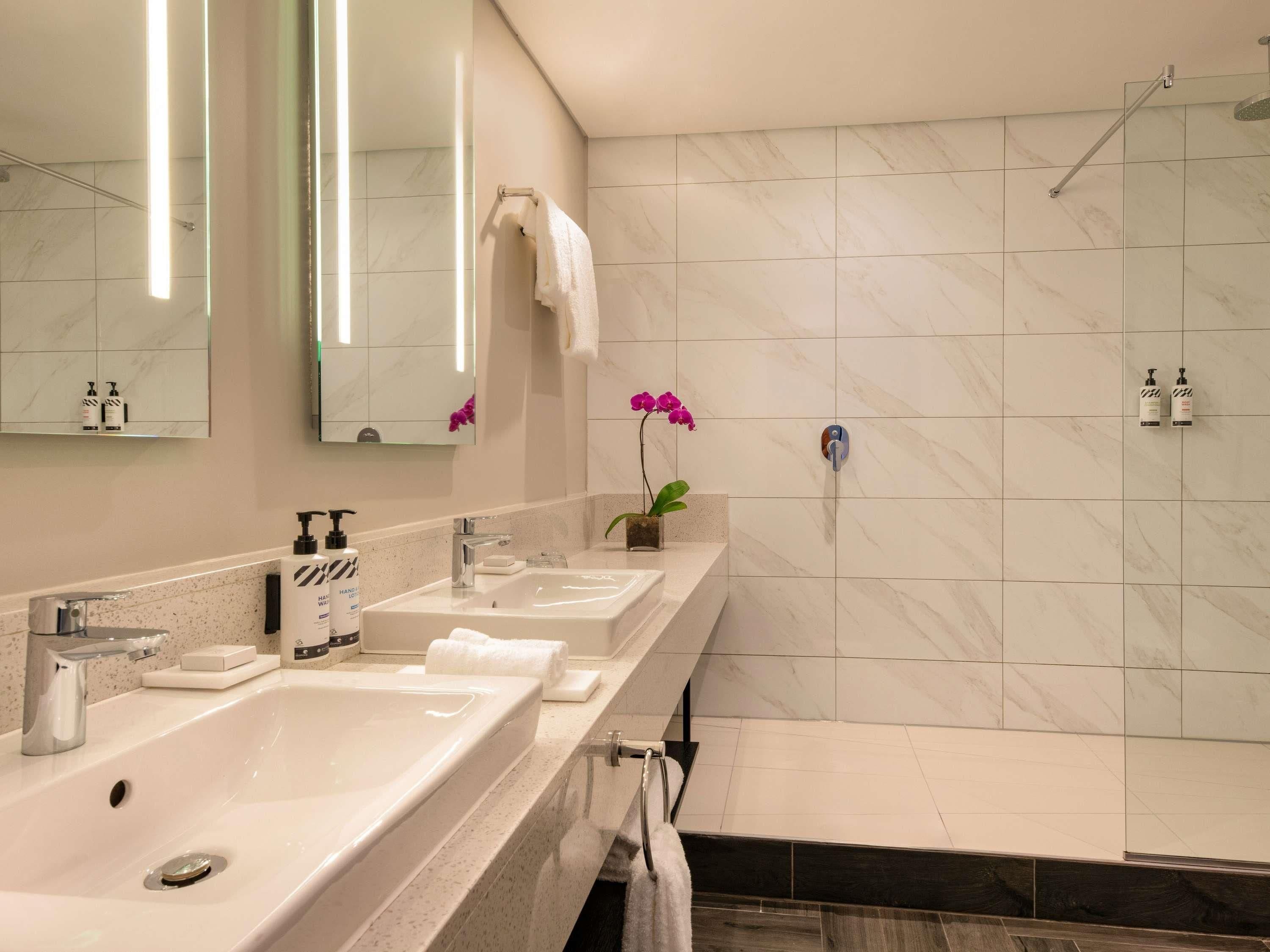 Un baño moderno con un espejo grande, dos lavamanos, una ducha y una orquídea en maceta.