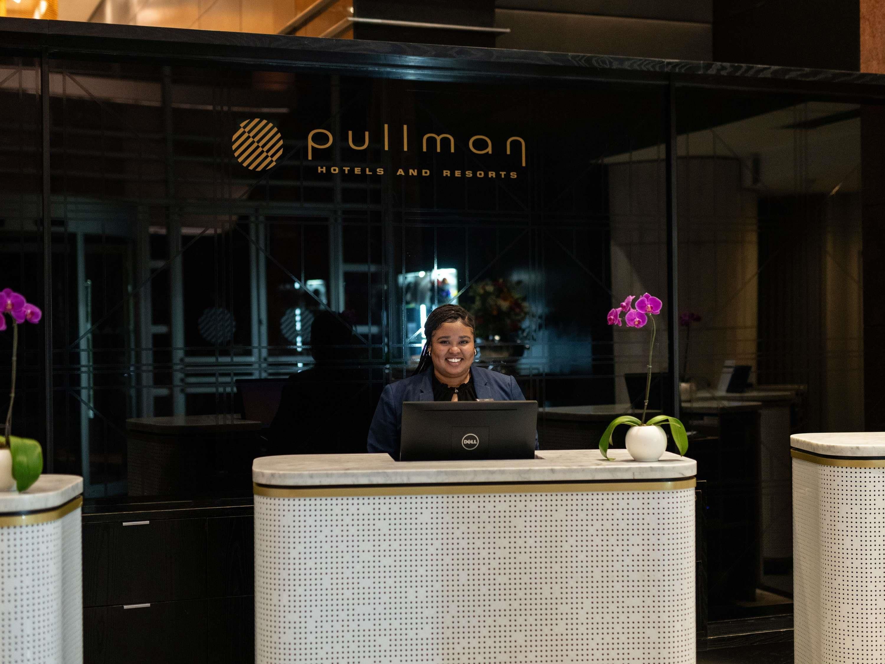 Un mostrador de recepción con una computadora portátil, un letrero de Pullman Hotels and Resorts y dos macetas con flores moradas.