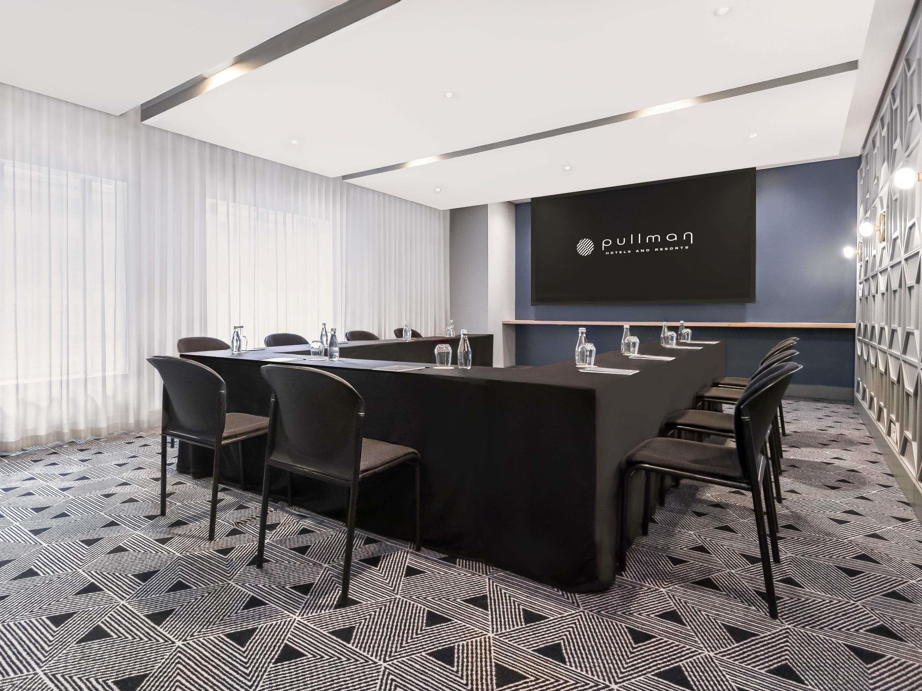 Ein Konferenzraum mit einem langen Tisch, Stühlen und einem Bildschirm, der das Logo 'Pullman Hotels and Resorts' zeigt.