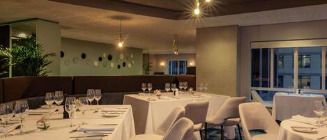 Un restaurante con varias mesas, cada una con copas de vino y cubiertos, y una lámpara de araña moderna.