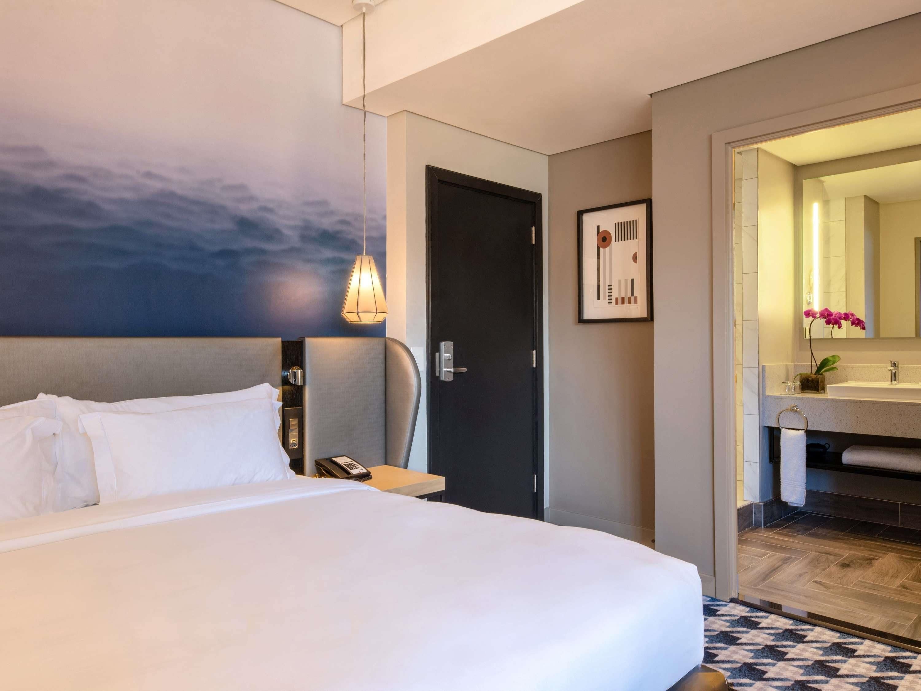 A hotel room with a bed, a desk, a phone, a door, a bathroom with a sink and mirror, and a wall with a painting.