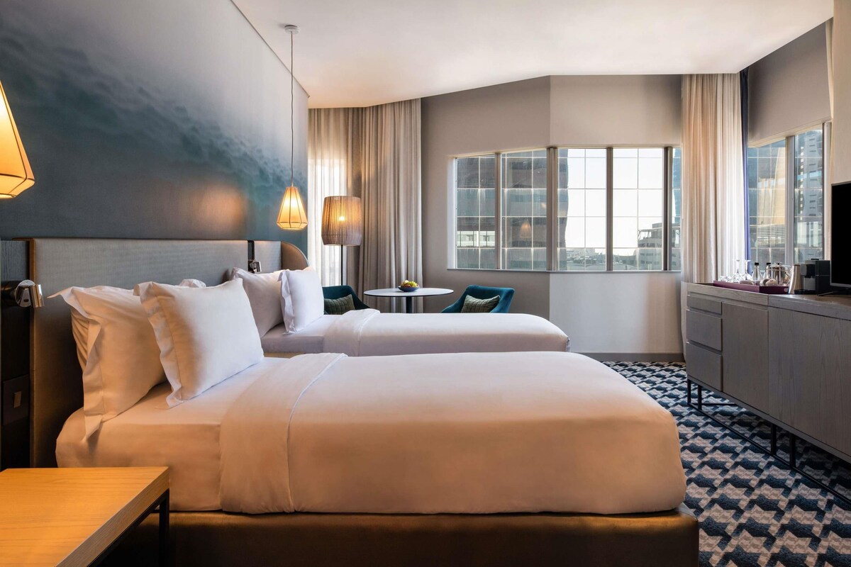 Quarto de hotel com duas camas, uma escrivaninha, uma mesinha, uma televisão e vista para a cidade.