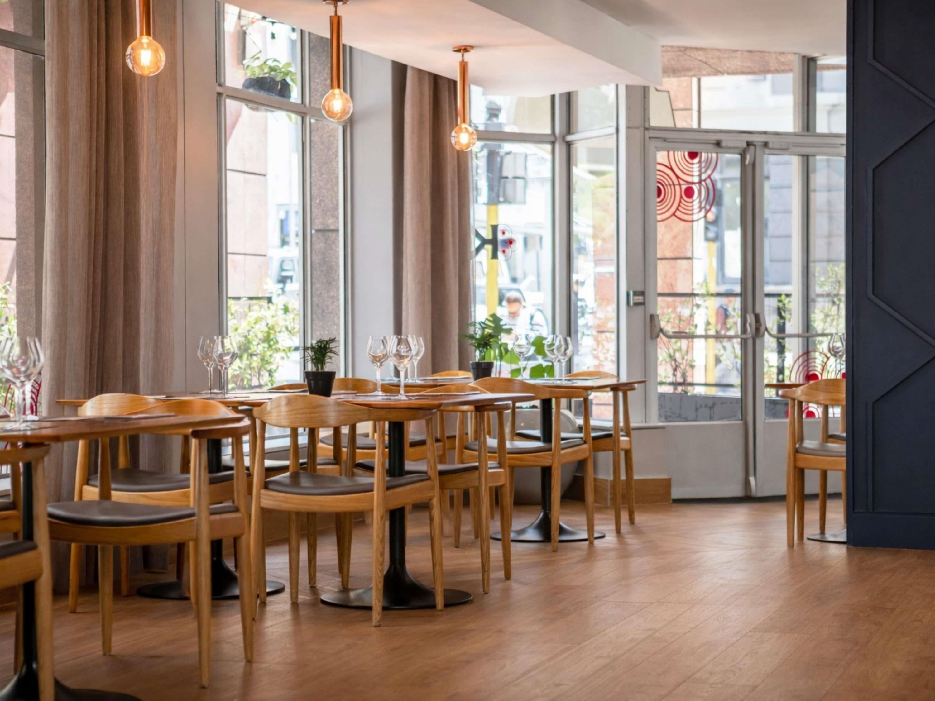 A restaurant interior with wooden tables and chairs, hanging pendant lights, and large windows with views of the street.