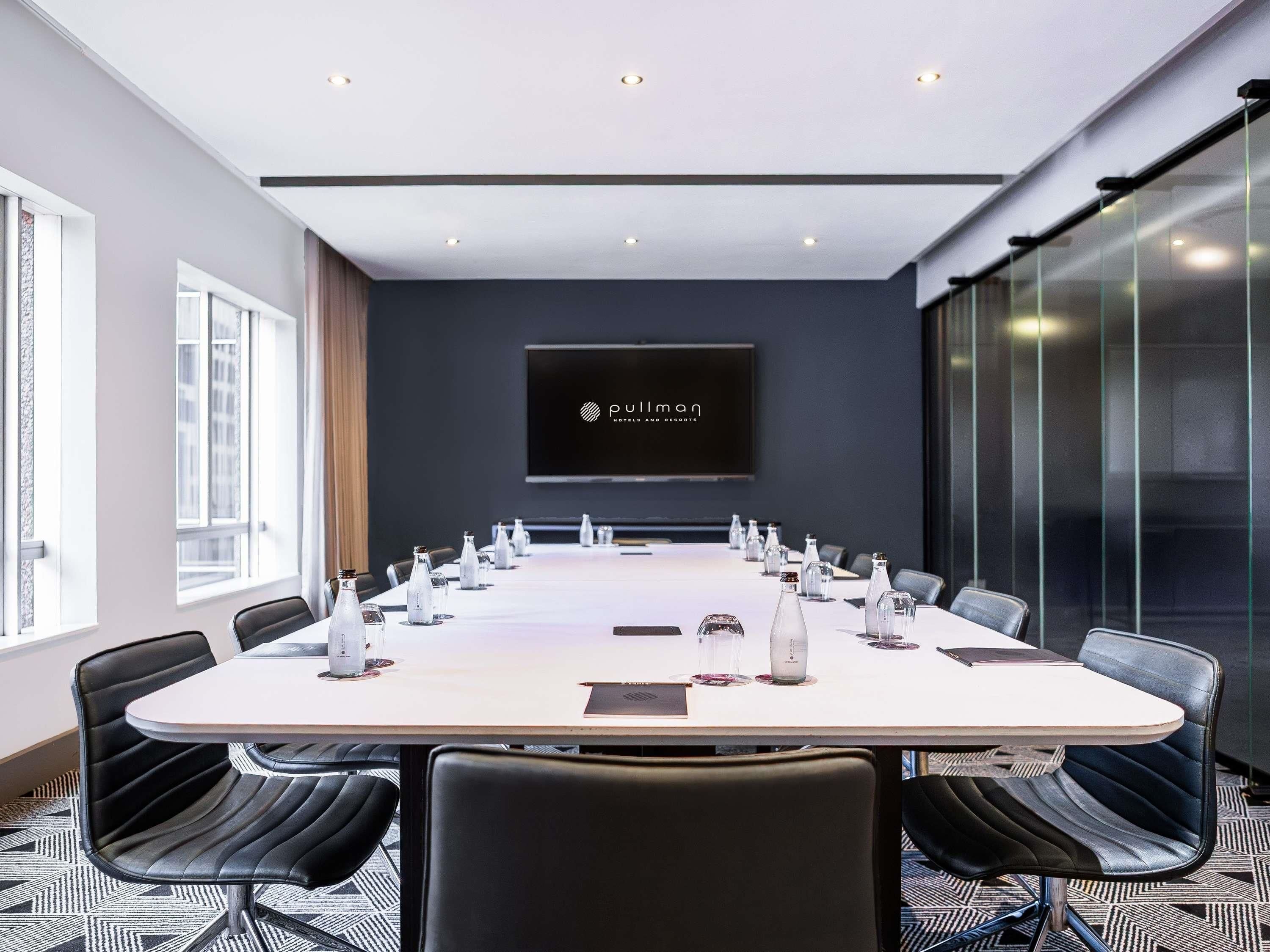 Un salón de conferencias con una larga mesa, sillas de cuero negro, un televisor de pantalla plana y un muro de cristal.