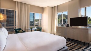Habitación de hotel con cama, escritorio, televisor y ventanas grandes con vista a la ciudad.