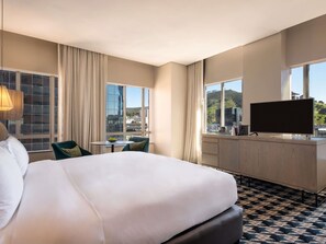 Habitación de hotel con cama, escritorio, televisor y ventanas grandes con vista a la ciudad.