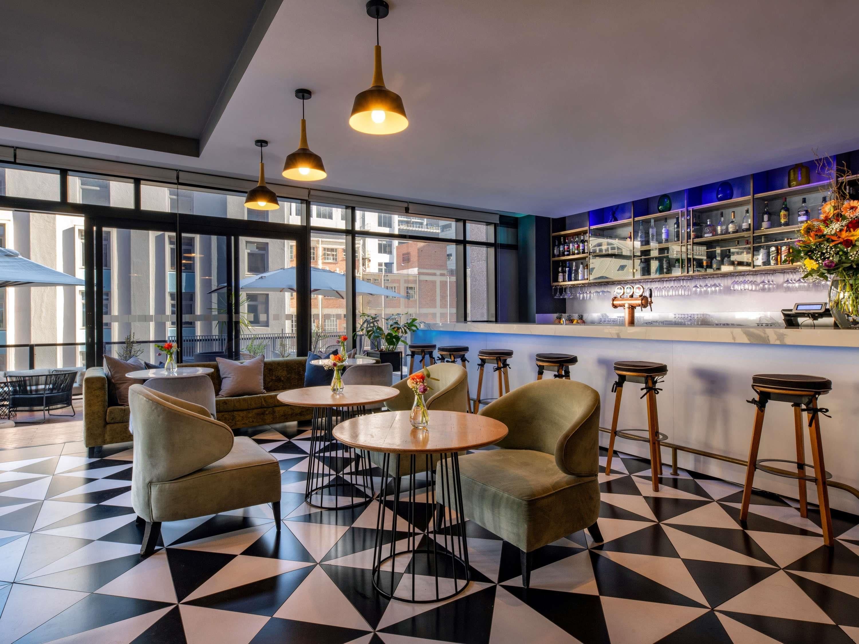 Un espace bar avec des tables rondes et des tabourets, un comptoir avec une caisse enregistreuse, et une vue sur la ville grâce à de grandes fenêtres.