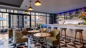 Un espace bar avec des tables rondes et des tabourets, un comptoir avec une caisse enregistreuse, et une vue sur la ville grâce à de grandes fenêtres.