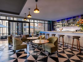 A bar area with round tables and bar stools, a bar counter with a cash register, and a view of the city through large windows.