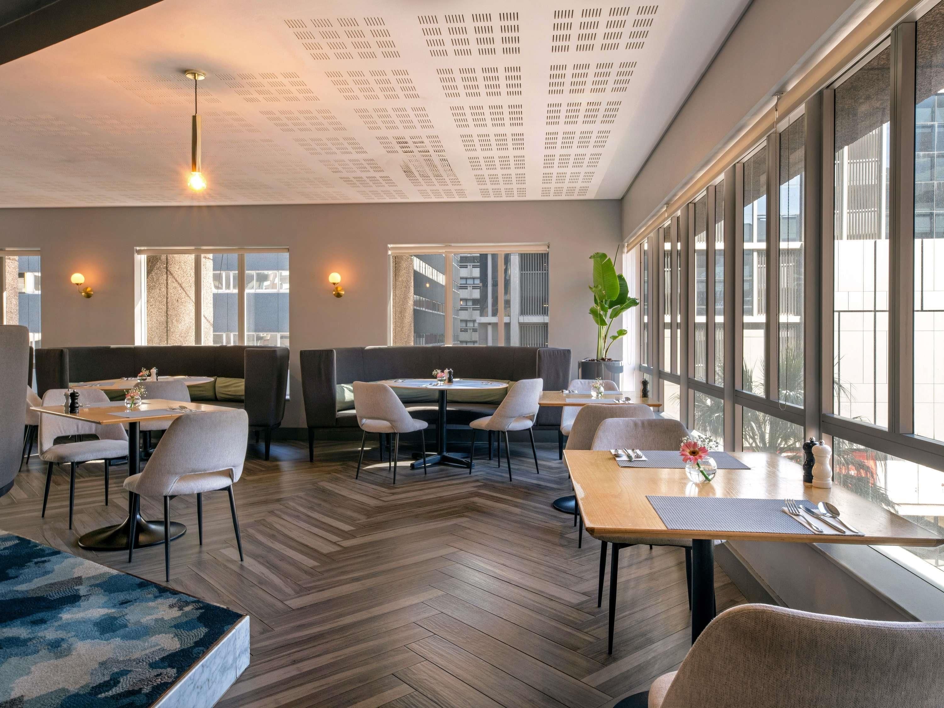 A modern dining area with wooden flooring, tables, chairs, and a large window providing a view of the city.