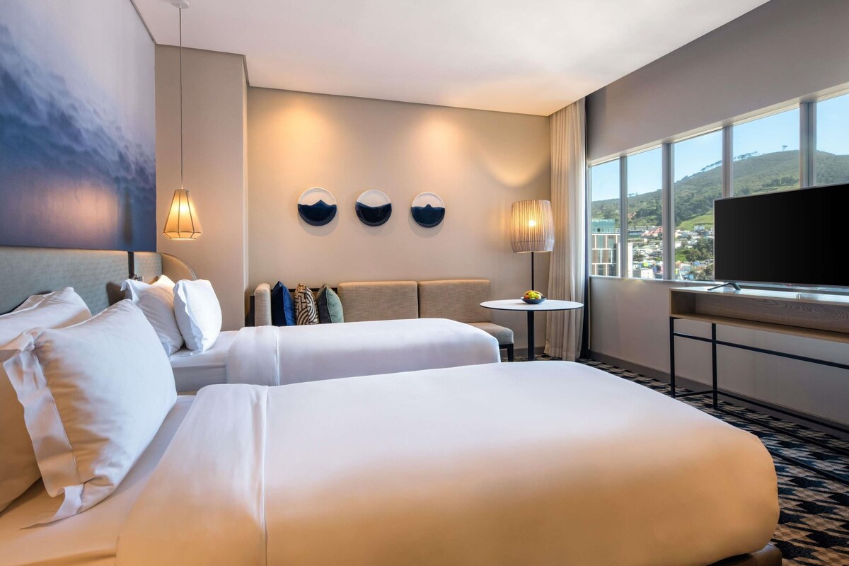 Quarto de hotel com duas camas, televisão, uma mesa pequena e vista para a paisagem urbana.
