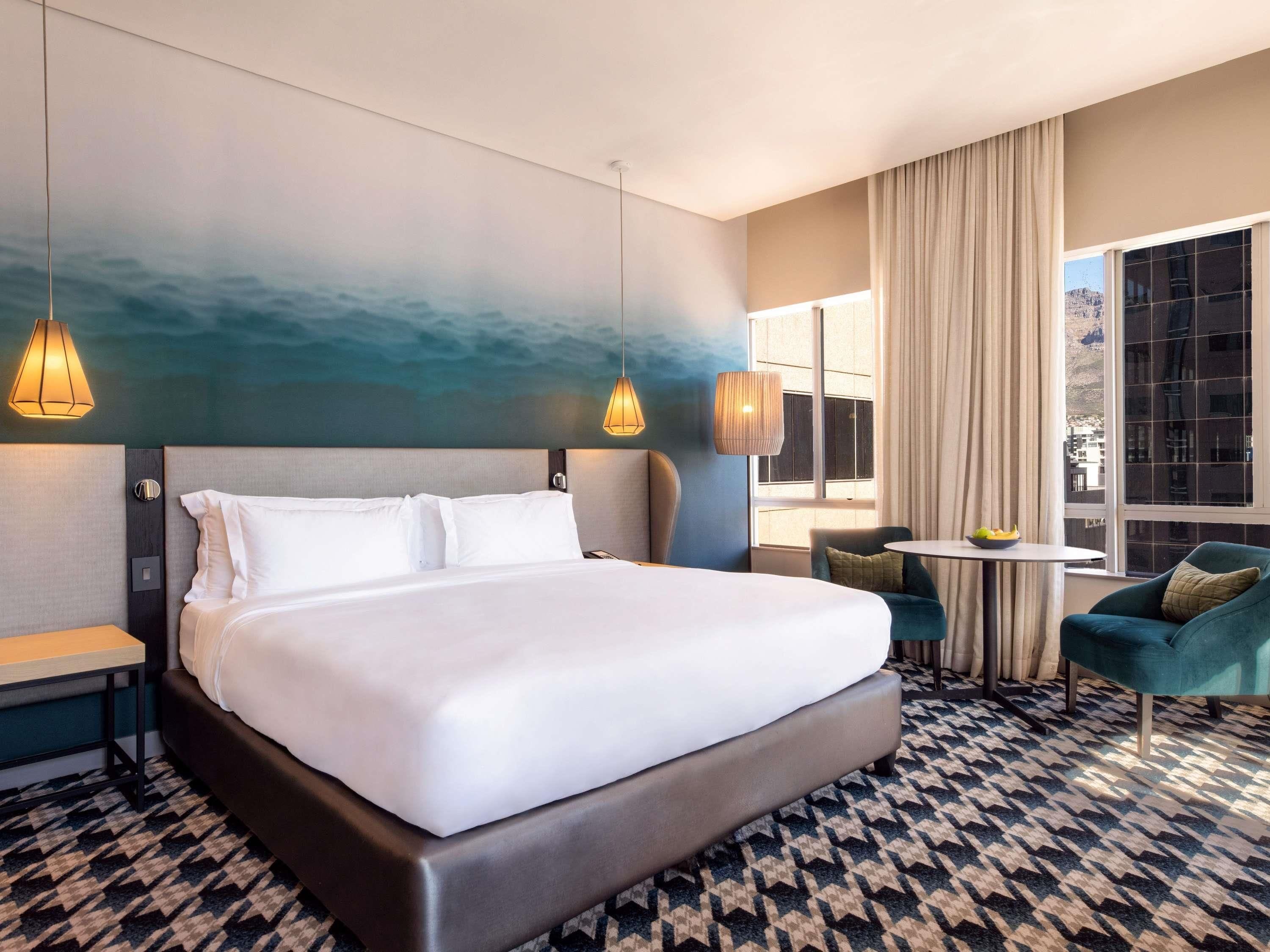 Habitación de hotel con una cama grande, dos mesitas de noche, una pequeña mesa redonda con un frutero, una silla y vistas a la ciudad a través de amplios ventanales.