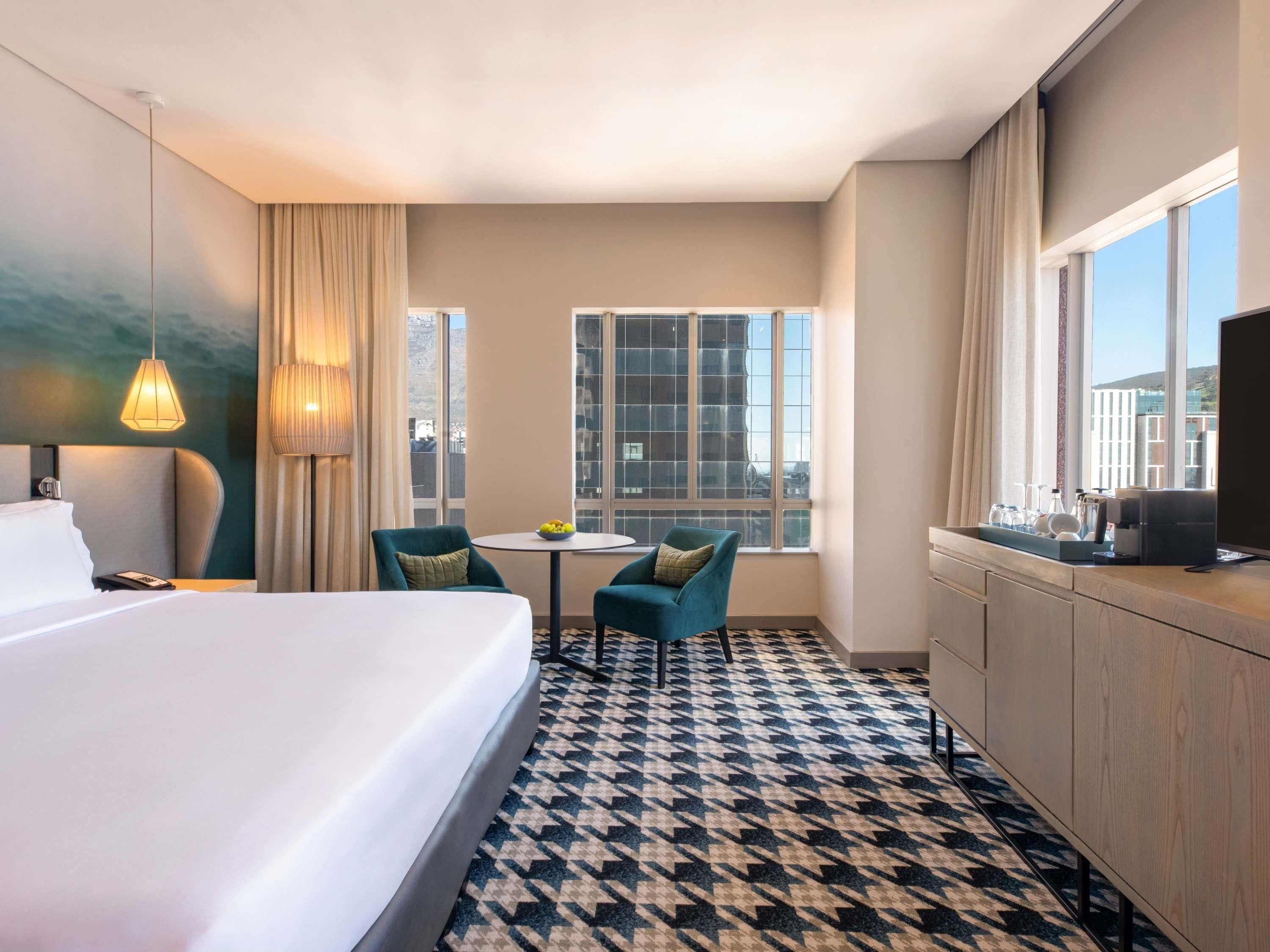 A hotel room with a bed, a desk, two chairs, a small table with a bowl of fruit, a television, and large windows.