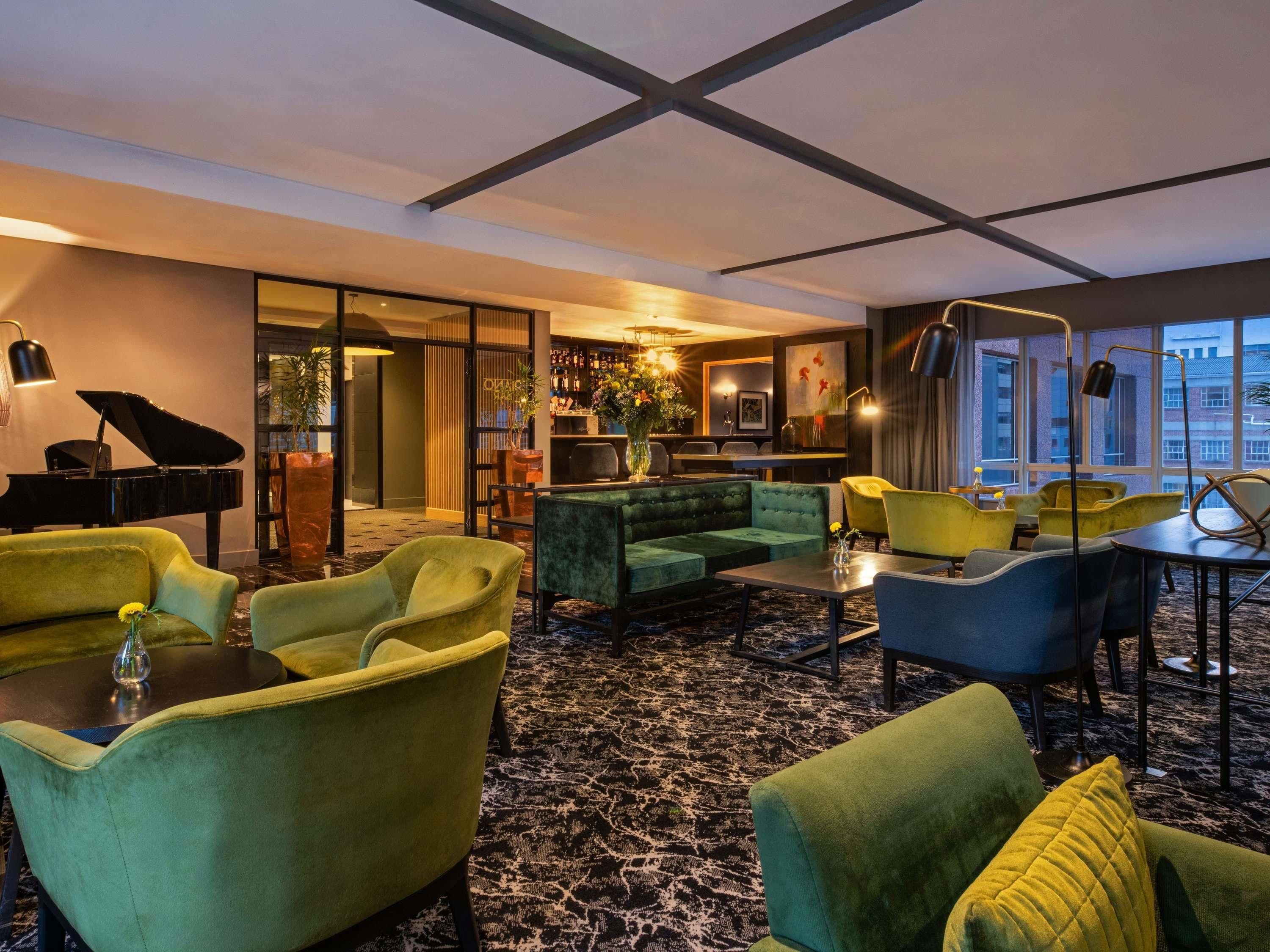 A modern hotel lobby with a grand piano, plush seating, a bar area, and large windows offering city views.